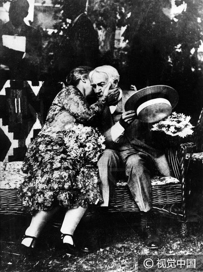 汤玛斯·爱迪生坐在长椅上，一位女子亲吻他的脸颊。